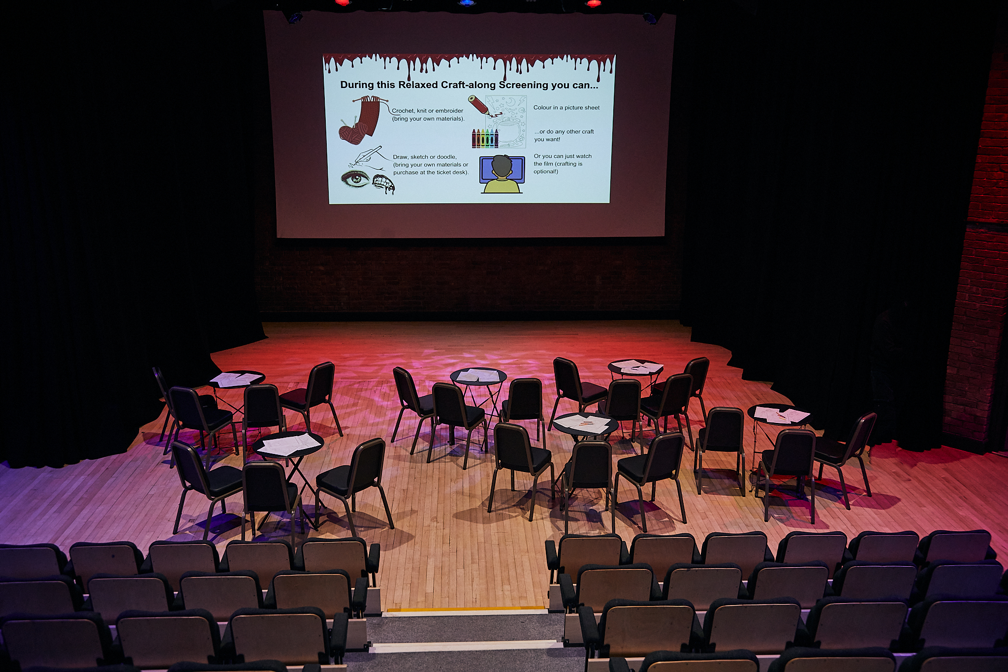 Photo of the cinema screen at Gosforth Civic Theatre, from the back. There are wooden chair backs visible and at the front space in front of the screen on the wood floor are tables and chairs with papers on the tables. On the screen is a slide which tells the audience what they can do during a Relaxed Craft-along screening.